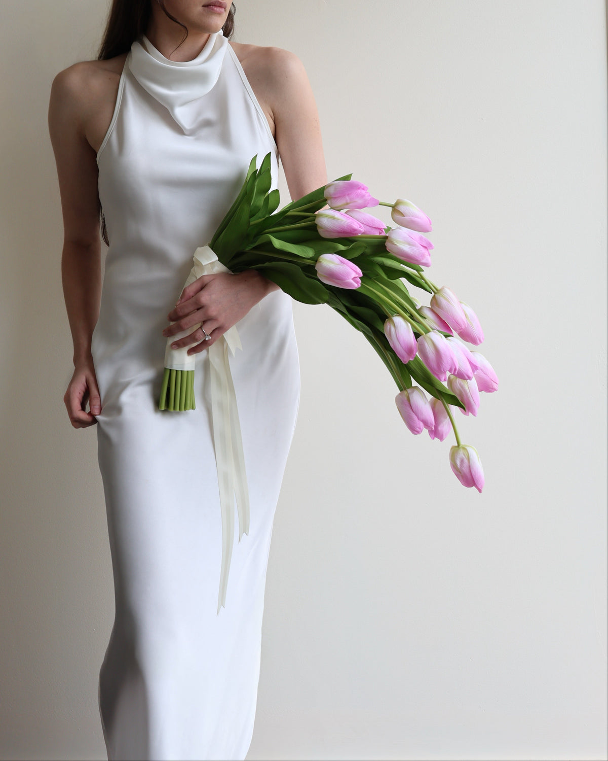 A woman wearing a sleeveless white cowl-neck dress stands against a plain light beige wall, holding a bouquet of pink tulips with green stems and white ribbons.