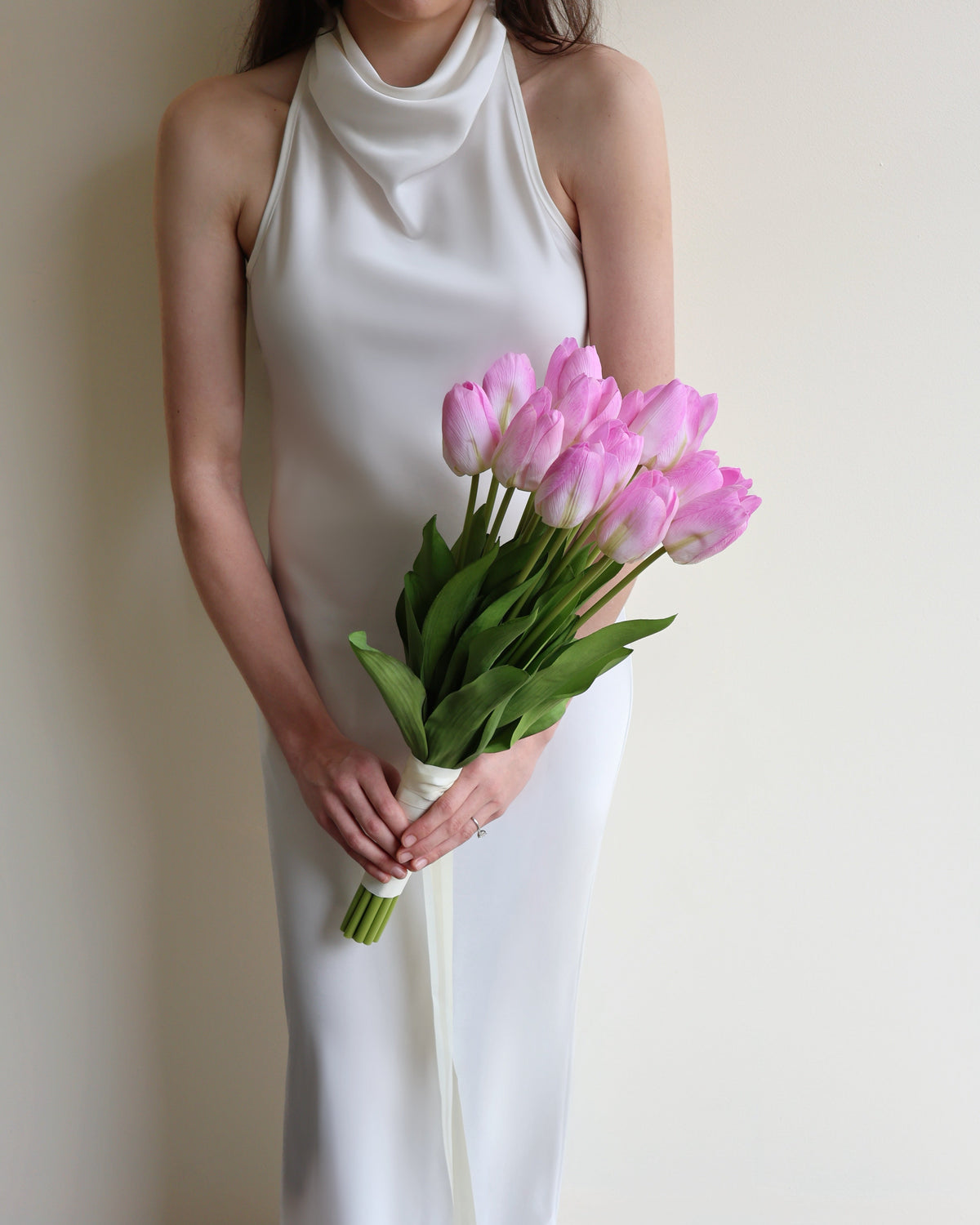 A woman wearing a sleeveless white cowl-neck dress holds a bouquet of pink tulips against a plain beige wall.