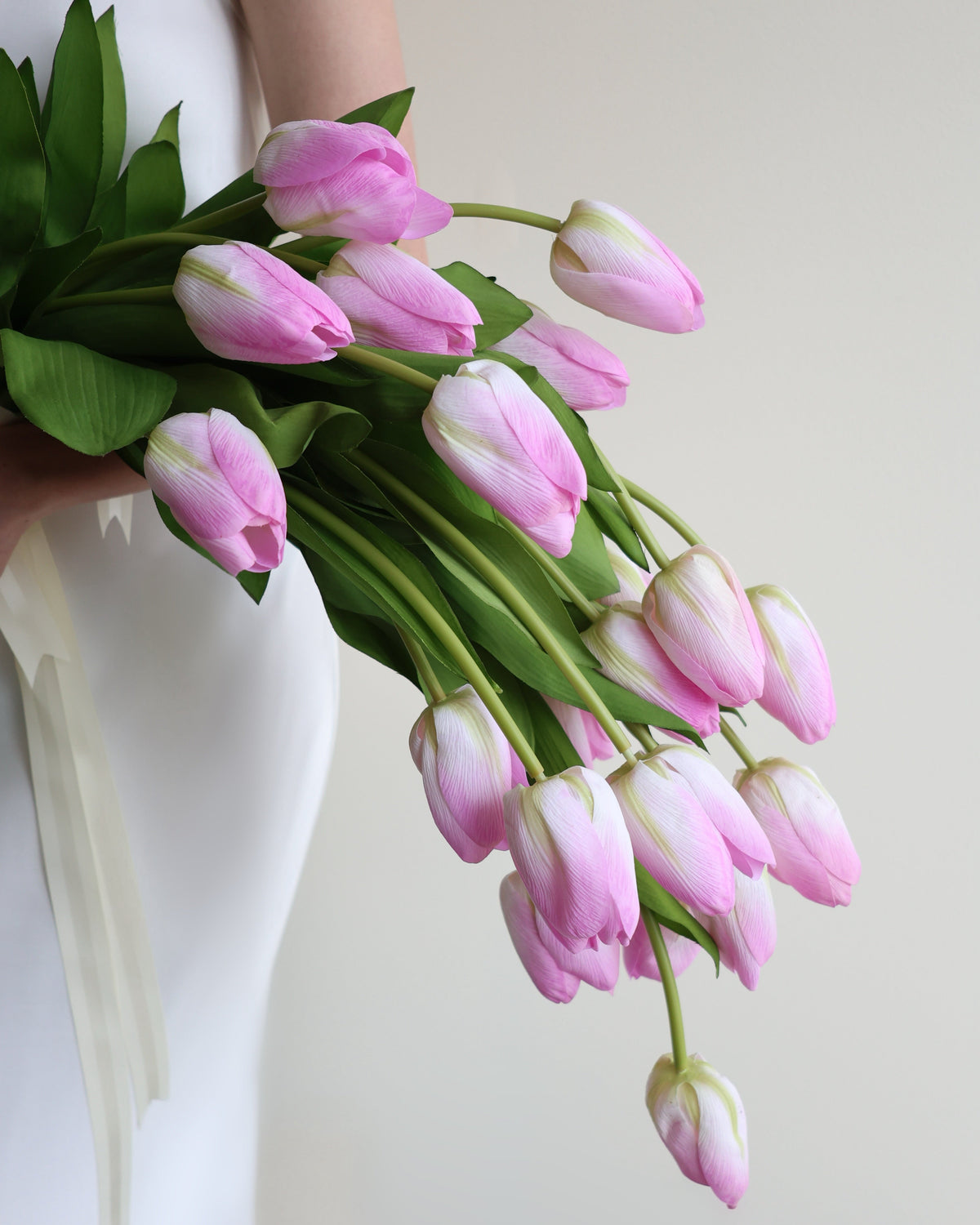 A bouquet of pink tulips with green leaves and stems, held by a person wearing a white sleeveless dress against a plain light background.