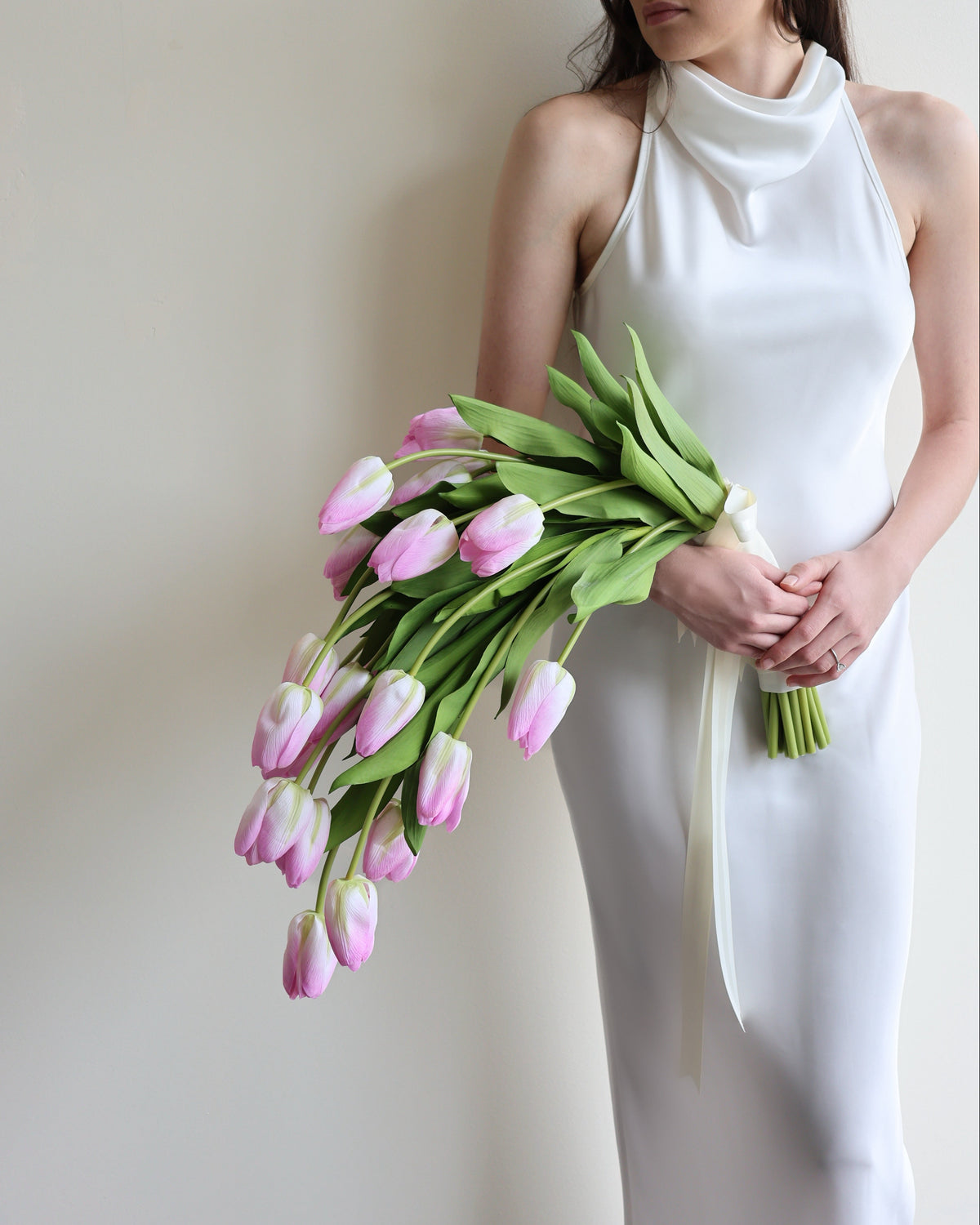A woman in a white sleeveless mock-neck dress holds a bouquet of pink tulips against a plain beige wall.